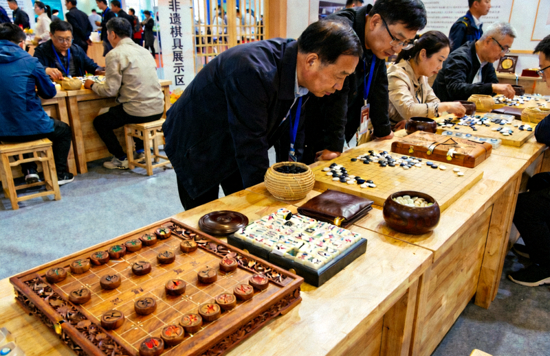 棋牌文化博览会现场，展台上陈列着传统非遗棋具，观众正在近距离观赏与体验